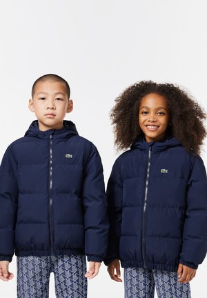 Giubbotti puffer navy con cappuccio, caratterizzati da un motivo a quadrati cucito e polsini elasticizzati. Logo Lacoste nella parte superiore sinistra di ciascun giubbotto.
