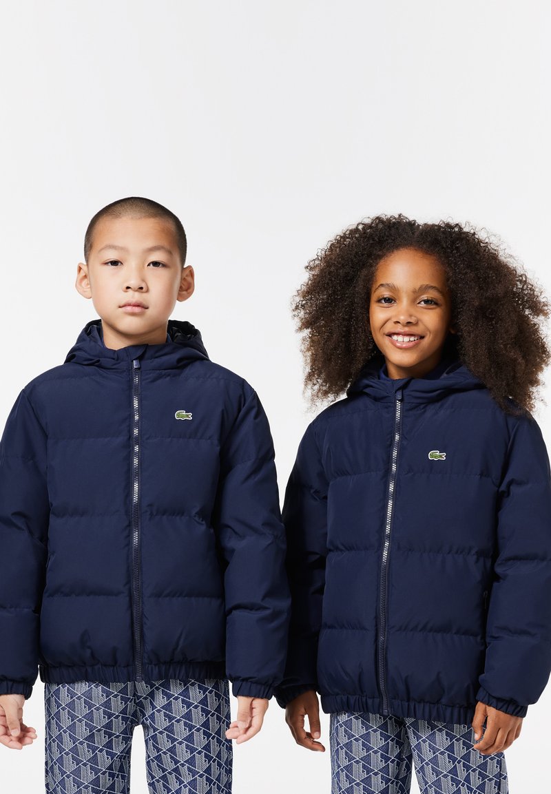 Vestes matelassées bleu marine avec capuches, présentant un motif matelassé et des poignets élastiques. Logo Lacoste en haut à gauche de chaque veste.
