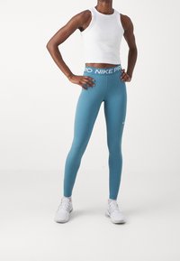Niebieskie dopasowane legginsy z szerokim pasem w talii z napisem "NIKE PRO" w białym kolorze. W połączeniu z białym bezrękawnikiem typu crop top i białymi butami sportowymi.