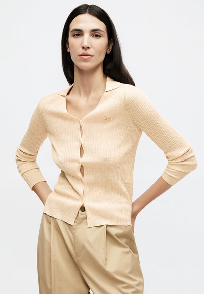 Γυναίκα με μακριά σκούρα μαλλιά, φορώντας μπεζ ριμπ πλεκτή ζακέτα με έμβλημα λεοπάρδαλης και ψηλόμεσα μπεζ παντελόνια, με τα χέρια στη μέση.