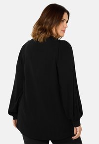 Blusa nera con maniche lunghe e svolazzanti, polsini con bottoni e un tessuto morbido con una silhouette leggermente rilassata e orlo arrotondato.