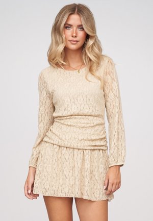 Jeune femme aux longs cheveux blonds portant une robe beige en dentelle à manches longues, debout devant un fond clair uni.