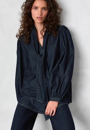 Frau mit lockigem Haar, die eine dunkle Denim-Bluse mit Puffärmeln und passende Hosen trägt, steht vor einem hellgrauen Hintergrund.