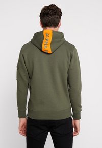 Felpa verde khaki con testo arancione "ALPHA" sulla schiena, dotata di cappuccio con cordino e tasca con zip sulla manica sinistra.