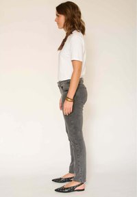 Femme portant une chemise blanche à manches courtes, un jean gris, des ballerines noires pointues avec œillets, une écharpe marron et des bracelets, debout de profil sur un fond uni.