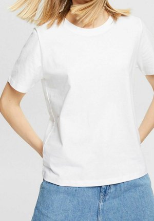 Kobieta ubrana w prosty biały t-shirt z krótkim rękawem i jasnoniebieskie dżinsy, ręce opierające się na biodrach, włosy lekko uniesione.