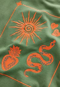 Zelená látka s výraznou oranžovou výšivkou slnka, hada, jahody a sŕdc, s detailnými čiarami a textúrami.