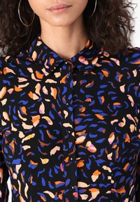 Black blouse with a collar, featuring an abstract pattern in orange, blue, and beige. Includes black buttons and a smooth fabric texture.