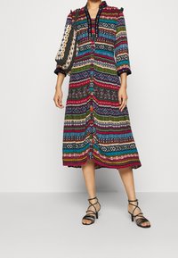 Robe à motifs multicolores avec un boutonnage sur le devant, des manches trois-quarts et des accentuations à pompons. Associée à des sandales noires à lanières.