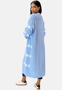 Largo cárdigan de punto azul con patrones de estrellas blancas, puños y dobladillo acanalados. Corte holgado, tejido con textura, diseño de frente abierto.