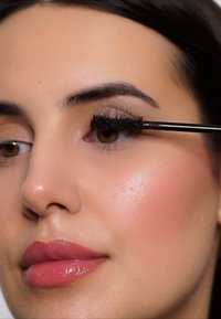 Schwarze Wimperntusche wird mit einem schwarzen Applikator auf die Wimpern aufgetragen. Die Haut hat eine glatte Textur mit einem subtilen Glanz. Die Lippen sind glänzend pink.