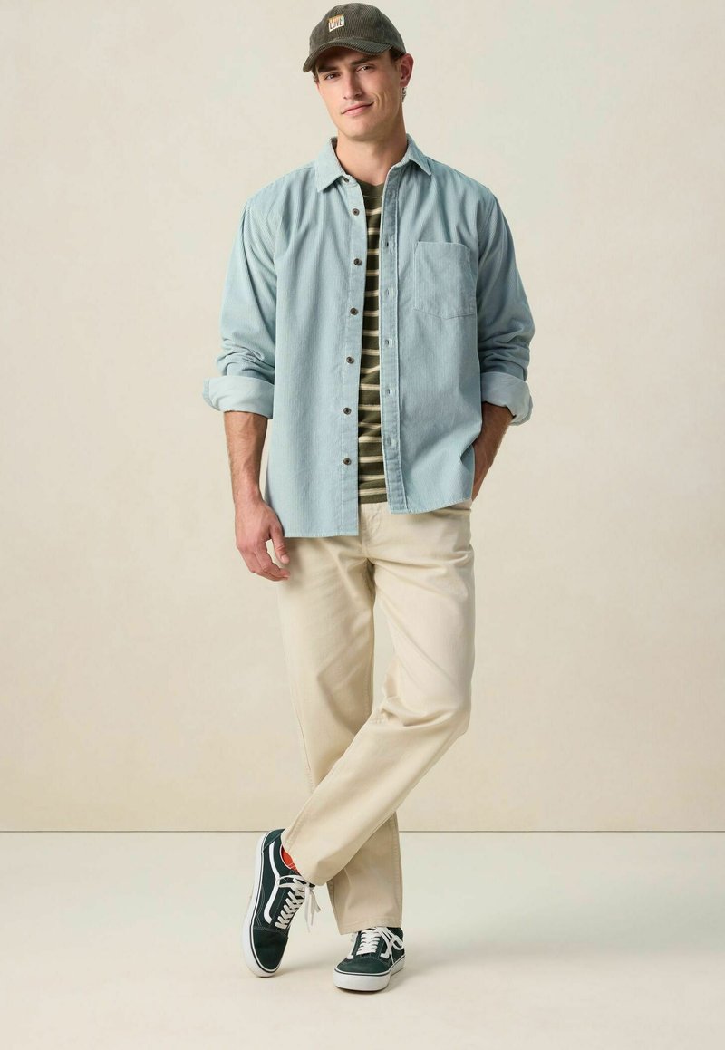 Chemise bleu clair à boutons par-dessus un T-shirt rayé vert et noir, pantalon beige et baskets navy avec semelles blanches. Casquette marron sur la tête.