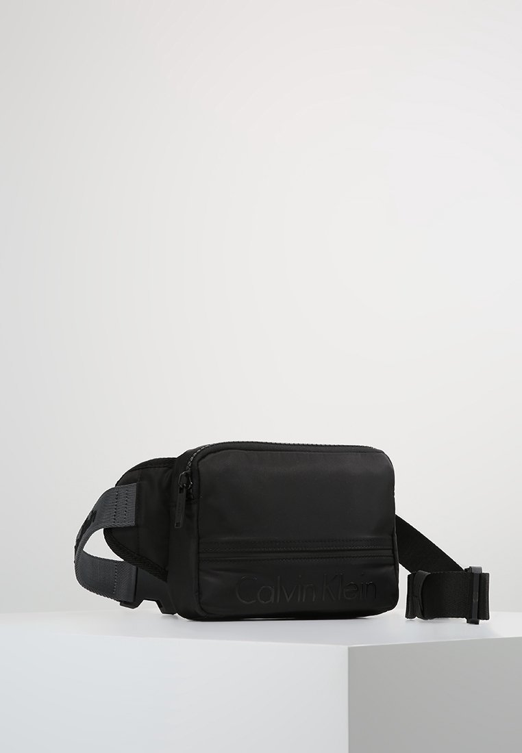 Schwarze Nylon-Crossbody-Tasche mit Reißverschluss, verstellbarem Tragegurt und eingeprägtem Logo auf der Vorderseite. Rechteckige Form mit glatter Textur.