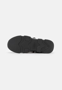 Semelle de sneaker noire en matériau caoutchouteux texturé. Présente un motif en zigzag, une pointe arrondie et un logo de la marque embossé près du talon.
