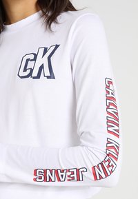Weißes Langarmhemd aus Baumwolle, mit navy blauem "CK"-Logo auf der Brust und rotem Text "CALVIN KLEIN JEANS" entlang des linken Ärmels.