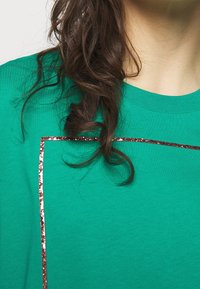 Lockar av brunt hår faller över en grön skjorta med en röd glittrig fyrkantig kontur nära halsringningen.