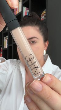 Transparente Tube des NARS Concealers mit schwarzem Deckel, der ein hellbeiges Produkt im Inneren zeigt, gehalten von einer Hand mit natürlichen Nägeln.