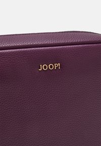 Sac en cuir violet avec une finition texturée, présentant un logo doré "JOOP!". Le design inclut une forme élégante et des accents minimalistes.