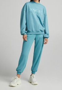 Turkos sweatshirt och sweatpants-set tillverkat av mjukt tyg, med rund hals och ordet "ANGEL" i vit tryck. Vita sneakers fulländar looken.