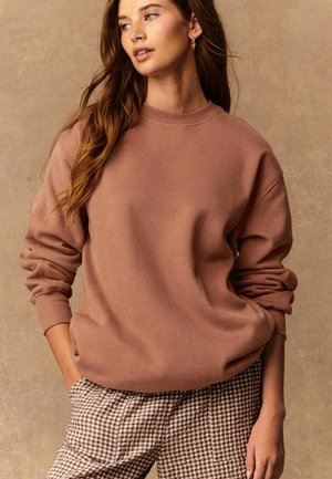 Sweatshirt surdimensionné marron avec des poignets côtelés, coupe décontractée et texture douce. Associé à un pantalon à carreaux marron et blanc.