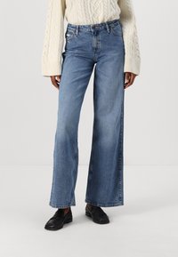 Mujer vestida con vaqueros de pierna ancha azul claro, suéter de punto de cable crema y mocasines negros, de pie contra un fondo blanco.