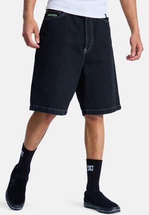 Άνδρας που φοράει μαύρα σορτς μέχρι το γόνατο, μαύρες κάλτσες DC, μαύρα παπούτσια χωρίς κορδόνια και λευκό πουκάμισο σε λευκό φόντο.