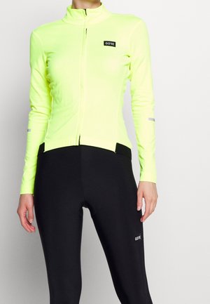 Neon gele lange mouw fietsjack met een rits aan de voorkant, zwarte accenten, een getailleerd ontwerp en reflecterende details op de mouwen, gecombineerd met zwarte leggings.