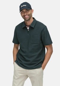 Mörkgrön pikétröja med halvdragkedja, korta ärmar och bröstficka, bärs med ljusa byxor och en marinblå keps.