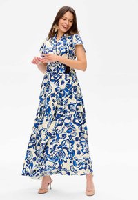 Robe maxi en crème avec motif floral bleu, manches courtes, décolleté en V et un design enveloppant accentué par une ceinture à la taille.
