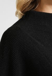 Black ribbed knit sweater with a high neck, featuring textured lines and a soft appearance. Details include a modern, minimalist design.