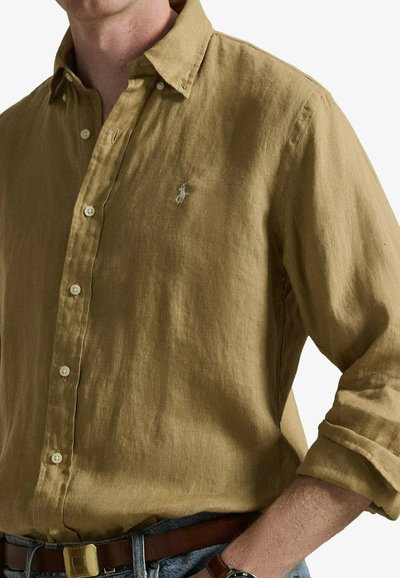 Homme portant une chemise en lin vert olive à boutons avec manches retroussées, ceinture marron, jean bleu et une montre noire au poignet gauche.