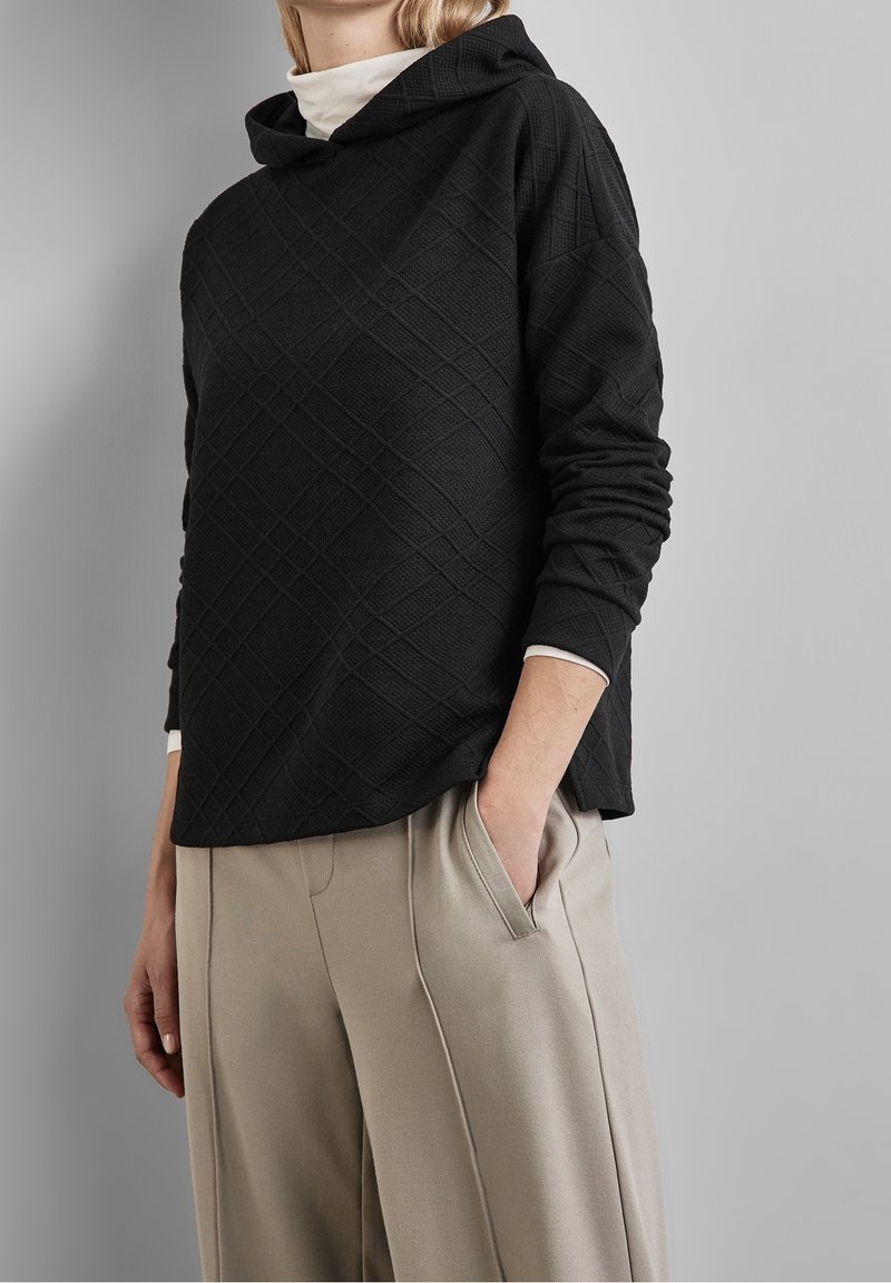 Sweat-shirt à capuche noir avec un motif en losanges texturé, présentant une coupe ample, des manches longues et des poches latérales, associé à un pantalon beige.