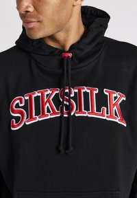 Bărbat îmbrăcat într-o hanorac negru cu logo-ul roșu și alb "SIKSILK" și șnururi negre cu stopere roșii, pe fundal alb.
