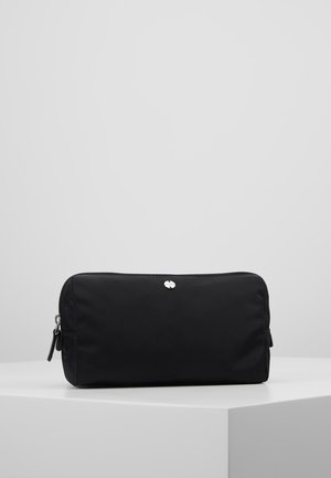Trousse de toilette rectangulaire noire avec une fermeture éclair sur le dessus et un petit logo rond argenté à l'avant, posée sur une surface blanche.