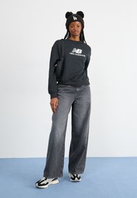 Fekete kapucnis pulóver fehér logóval, szürke bővülő farmernadrág és fekete-fehér sportcipő. Kiegészíti egy zöld kötött sapka pom-pomokkal.