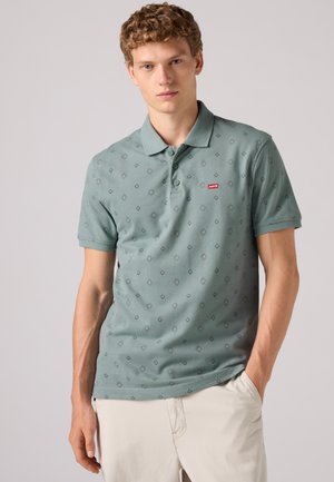 Ung mand med krøllet hår iført en lysegrøn Levi's polo med mønster og lyse beige bukser med den ene hånd i lommen.