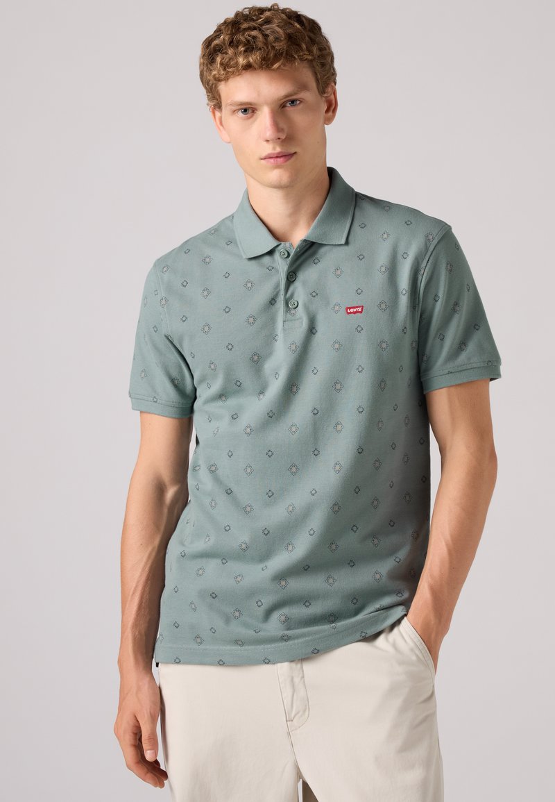 Jeune homme aux cheveux bouclés portant un polo Levi's vert clair à motifs et un pantalon beige clair, une main dans la poche.