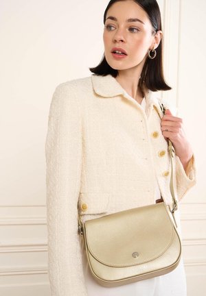 Femme aux cheveux foncés portant une veste texturée crème avec des boutons dorés, portant un sac à bandoulière en cuir doré clair devant un mur couleur crème.