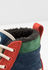 Baskets montantes en daim avec un assortiment de couleurs bleu, vert, rouge et crème. Dotées d'une doublure noire texturée et d'une languette de tirage rouge au talon.