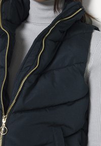 Czarna pikowana kamizelka z wysokim kołnierzem, wyposażona w złoty zamek błyskawiczny i boczne kieszenie. Nosi się ją na jasnoszarym prążkowanym golfie.