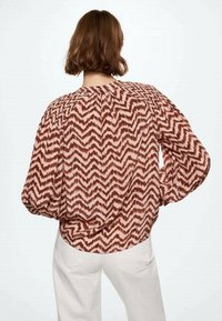 Donna con capelli corti castani che indossa una blusa ampia a maniche lunghe con motivi a zigzag marroni e rosa e pantaloni bianchi, in piedi con una mano sui fianchi.