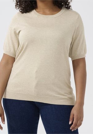 Woman wearing a beige short-sleeve knit top paired with dark blue jeans against a plain light background.