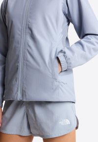 Άτομο που φοράει ανοιχτό μπλε αντιανεμικό μπουφάν με φερμουάρ και αντίστοιχα σορτς με το λογότυπο The North Face στο αριστερό πόδι.