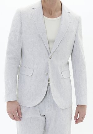 Homme portant une veste de costume à fines rayures gris clair et blanc avec un pantalon assorti sur une chemise blanche côtelée, debout devant un fond blanc.