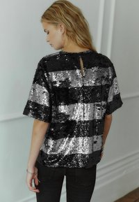 Top con paillettes in strisce nere e argento, con maniche ampie, colletto rotondo e dettaglio a forma di chiave nella parte posteriore. Finitura testurizzata con accenti riflettenti.