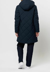 Persona con un abrigo acolchado largo con capucha de color azul marino, pantalones oscuros y zapatillas blancas, de pie con la espalda hacia el espectador sobre un fondo liso.