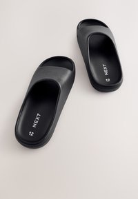 Schwarze Slide-Sandalen mit glatter, matter Oberfläche, offenem Zehendesign und einem niedrigen, gebogenen Absatz. Auf der Innensohle sind "NEXT" und die Größe "12" vermerkt.