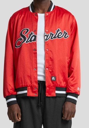 Personne portant une veste Starter rouge avec boutons-pression, des poignets rayés noir et blanc, et une chemise blanche côtelée en dessous.