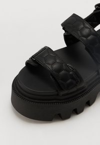 Black platform sandals with quilted texture, wide straps, and a rugged sole. Features a smooth inner surface and a rounded toe design.