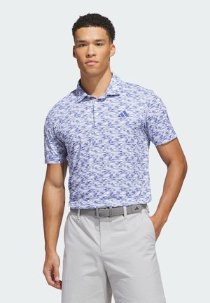 Mand iført en blå polo med mønster og lysegrå shorts, stående med den ene hånd i lommen mod en ensfarvet baggrund.